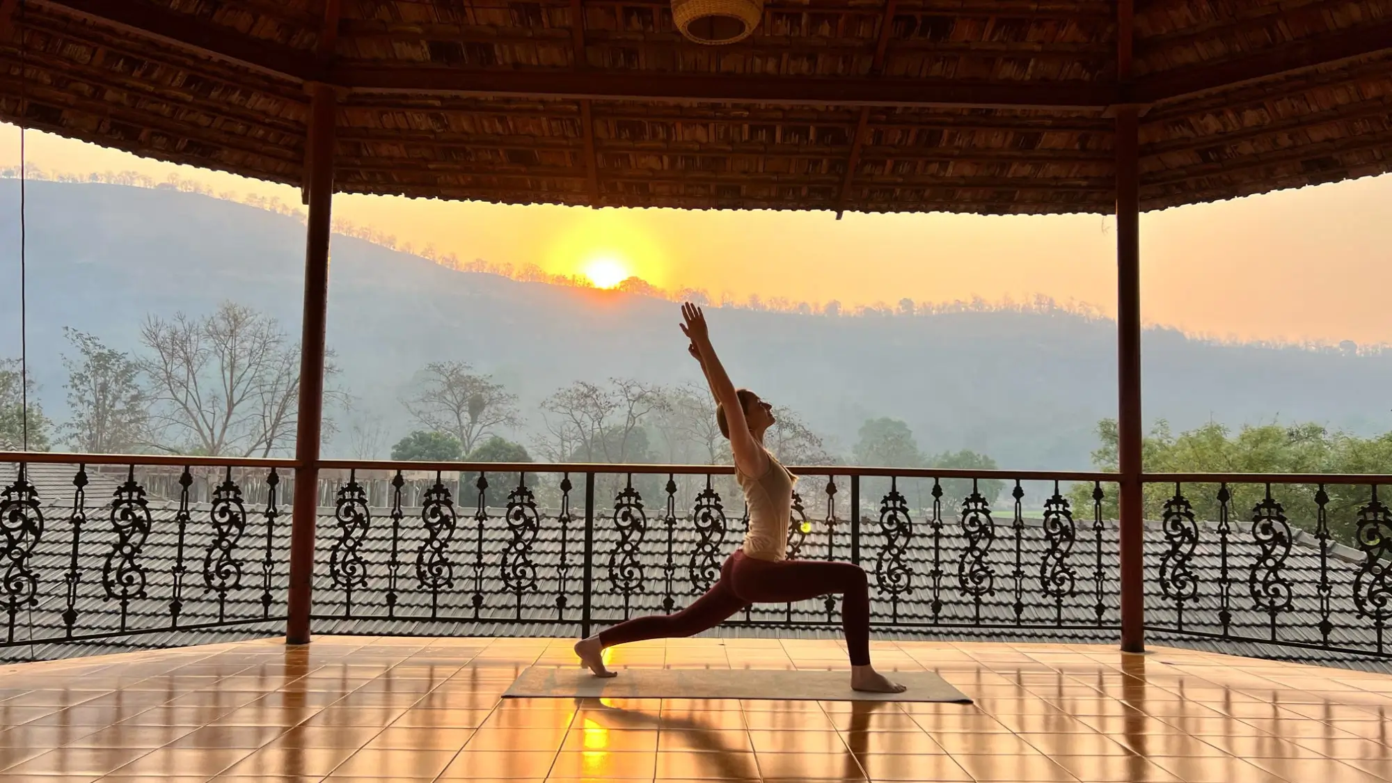 eine Frau steht in Krieger 1 Pose in der offenen Yoga Shala des Indien Ashrams, im Hintergrund ist der Sonnenaufgang über dem Govardhan Mountain zu sehen