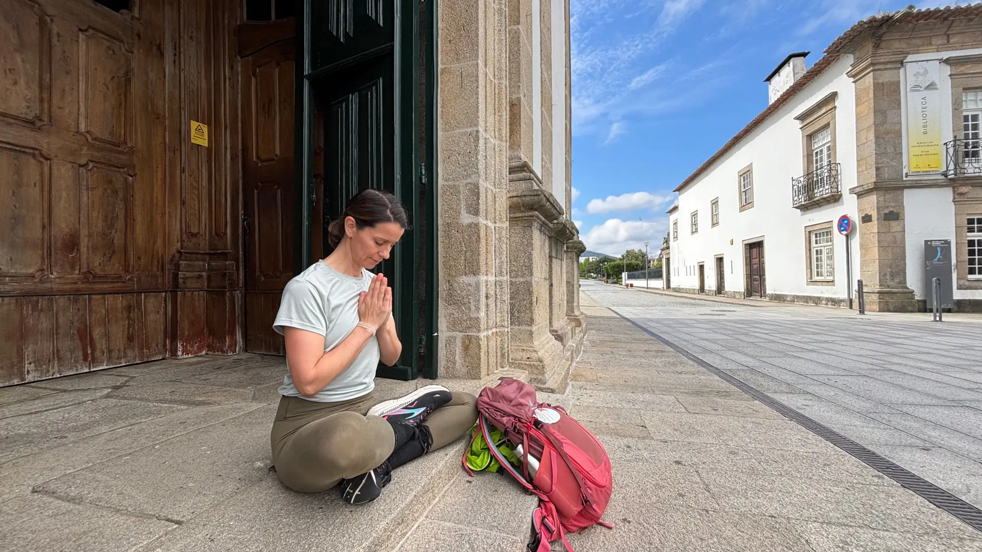 Eine Frau sitzt im Yogasitz und Kopf gesenkt vor einer Kirche, vor ihr steht der Wanderrucksack mit der Jakobsmuschel