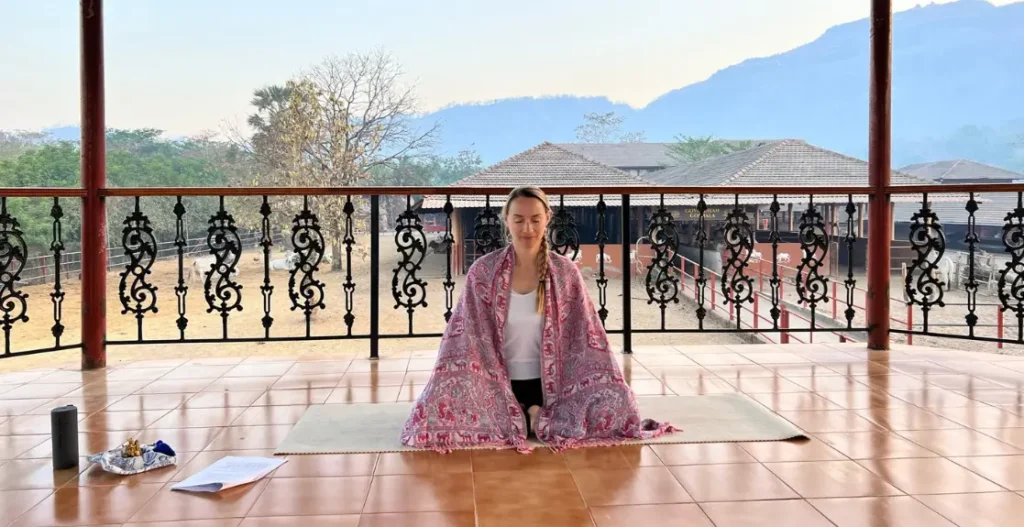 Frau sitzt im Yogasitz in einer Yoga Shala in Indien mit den heiligen Kuhställen und den Bergen nördlich von Mumbai im Hintergrund.