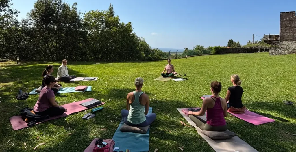 7 Frauen sitzen im Garten im Kreis auf ihren Yogamatten und im Hintergrund ist das Meer zu sehen
