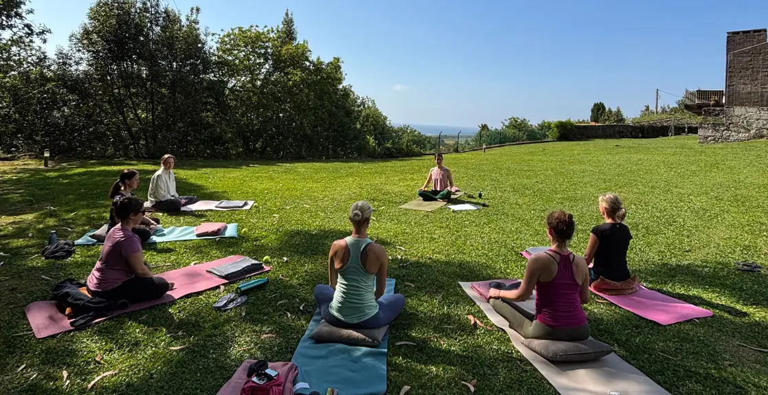 7 Frauen sitzen im Garten im Kreis auf ihren Yogamatten und im Hintergrund ist das Meer zu sehen